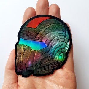 Pode incluir: Adesivo hologr&aacute;fico com um design de capacete futurista. O adesivo tem uma borda preta e um efeito de mudan&ccedil;a de cor, com tons de vermelho, verde, azul e dourado. O capacete tem uma viseira e detalhes intrincados, sugerindo um tema de fic&ccedil;&atilde;o cient&iacute;fica ou jogos.