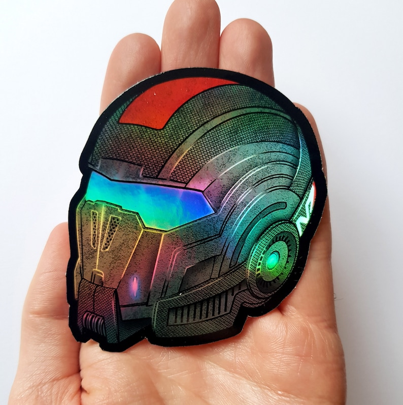 Pode incluir: Adesivo hologr&aacute;fico com um design de capacete futurista. O adesivo tem uma borda preta e um efeito de mudan&ccedil;a de cor, com tons de vermelho, verde, azul e dourado. O capacete tem uma viseira e detalhes intrincados, sugerindo um tema de fic&ccedil;&atilde;o cient&iacute;fica ou jogos.