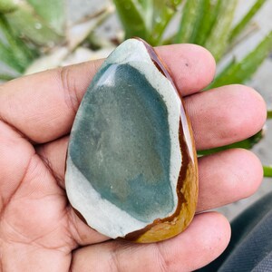 May include: A polished, teardrop-shaped gemstone with a mix of colors. The stone features a central area of muted teal, bordered by a white band and a brown edge. The overall shape is smooth and organic.
