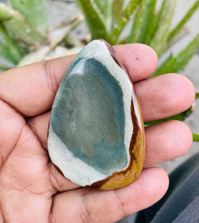 May include: A polished, teardrop-shaped gemstone with a mix of colors. The stone features a central area of muted teal, bordered by a white band and a brown edge. The overall shape is smooth and organic.