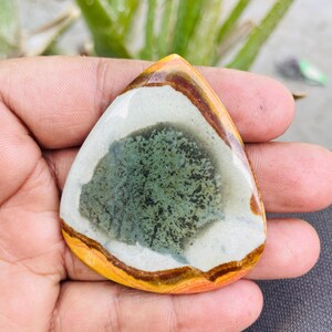 May include: A teardrop-shaped gemstone with a polished surface. The stone features a central area of mottled green and black, surrounded by a white band, a brown band, and a yellow-orange outer edge. The gemstone is held in a hand.