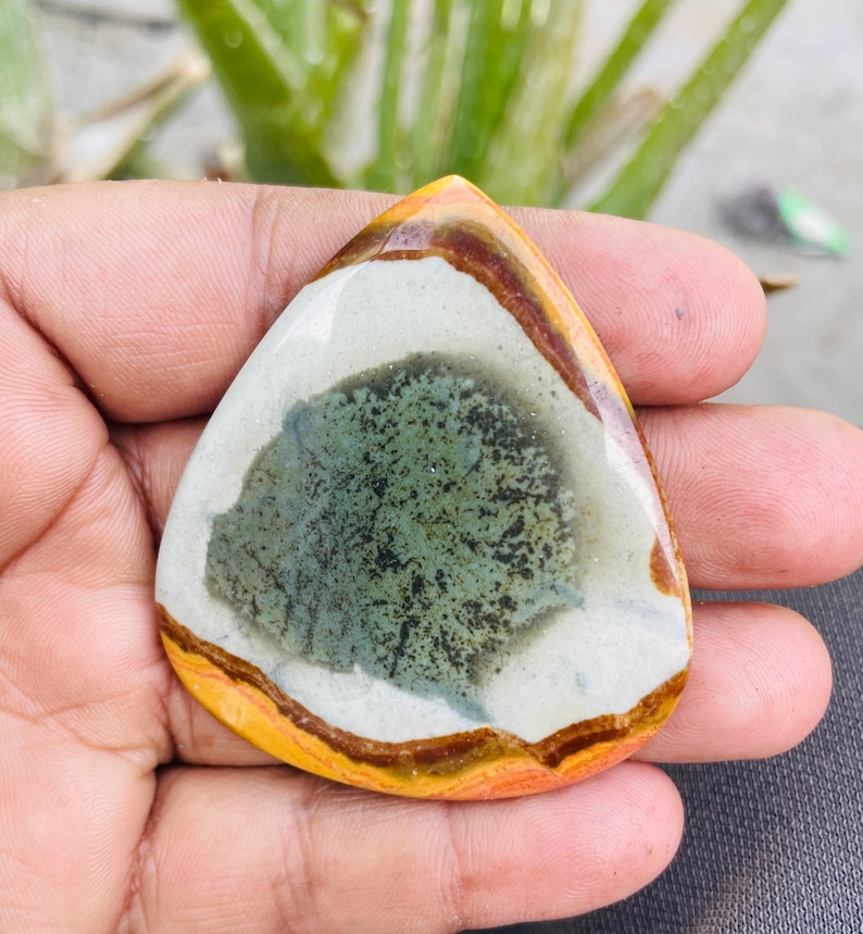 May include: A teardrop-shaped gemstone with a polished surface. The stone features a central area of mottled green and black, surrounded by a white band, a brown band, and a yellow-orange outer edge. The gemstone is held in a hand.