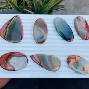 May include: Seven polished gemstones in various shapes and colours, including red, brown, green, and blue, are displayed on a white, ridged surface. The stones have unique patterns and textures, showcasing natural variations. The arrangement highlights the gemstones' beauty.