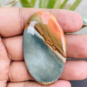 May include: An oval-shaped gemstone with a polished surface. The stone features a blend of colors, including shades of orange, brown, white, and gray. The gemstone is held in a hand, showcasing its natural beauty and unique color patterns.