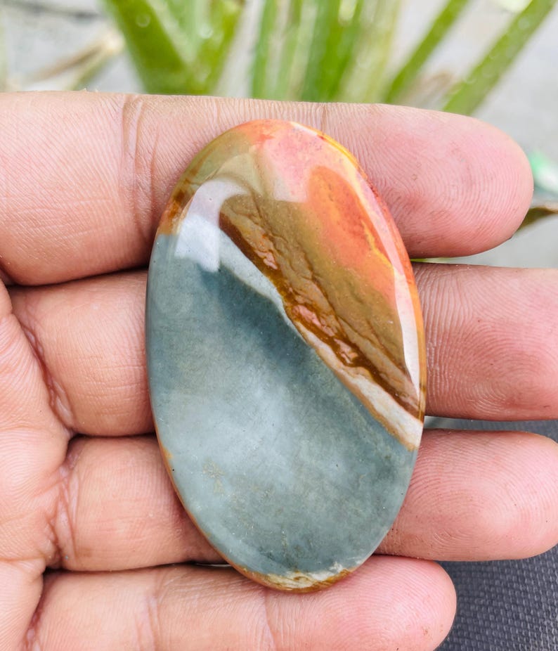 May include: An oval-shaped gemstone with a polished surface. The stone features a blend of colors, including shades of orange, brown, white, and gray. The gemstone is held in a hand, showcasing its natural beauty and unique color patterns.