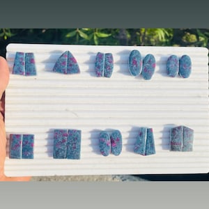 Pode incluir: Uma coleção de pedras preciosas de várias formas, incluindo retângulos e ovais. As pedras são uma mistura de tons azul-cinza e vermelho, dispostas sobre uma superfície branca e estriada. Provavelmente para criação de joias.