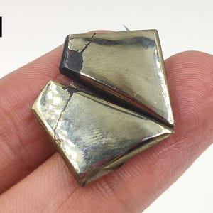 Pode incluir: Um objeto geométrico, com aspeto metálico, com um design dividido. O objeto tem uma superfície brilhante e refletora com um esquema de cores ouro e prata. O objeto é segurado pelo dedo de uma pessoa.