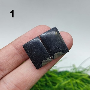Pode incluir: Duas pedras preciosas retangulares polidas, com uma cor cinza carvão escuro. As pedras têm uma superfície ligeiramente texturizada e um brilho metálico. As bordas inferiores das pedras têm uma aparência mais clara e manchada. O número "1" é visível no canto superior esquerdo.