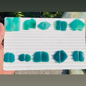 May include: A selection of polished, teal-coloured gemstone cabochons in various shapes, including rectangles, triangles, and rounded forms. The gemstones are displayed on a white, textured surface, showcasing their vibrant colour and smooth finish.
