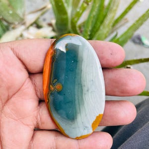 May include: Oval-shaped polished stone with a mix of colors. The stone features bands of orange, teal, and white, with a smooth, glossy surface. The stone is held in a hand, showcasing its natural beauty and unique patterns.