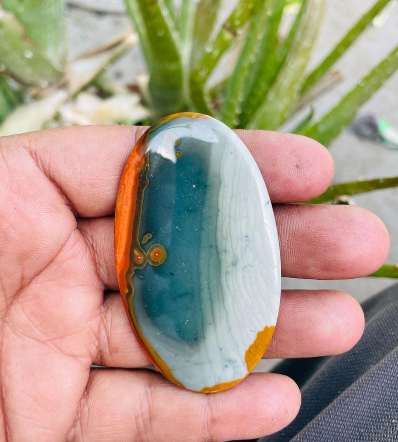 May include: Oval-shaped polished stone with a mix of colors. The stone features bands of orange, teal, and white, with a smooth, glossy surface. The stone is held in a hand, showcasing its natural beauty and unique patterns.