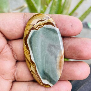 May include: An oval-shaped polished gemstone with a mix of colors. The stone features a central area of muted green, bordered by bands of brown, tan, and white. The gemstone is held in a hand, showcasing its natural patterns and textures.