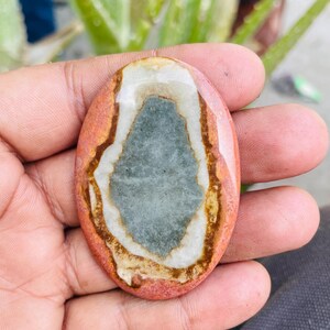 May include: Oval-shaped polished stone with a central gray-blue area, surrounded by white, brown, and reddish-orange bands. The stone is held in a hand, showcasing its natural colors and patterns. A unique, natural gemstone.
