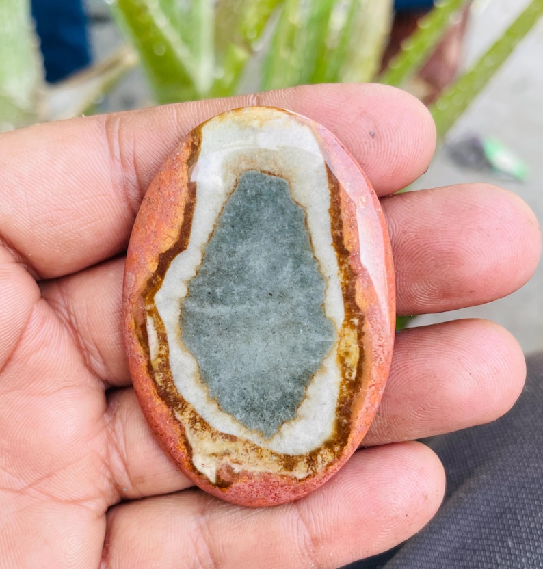 May include: Oval-shaped polished stone with a central gray-blue area, surrounded by white, brown, and reddish-orange bands. The stone is held in a hand, showcasing its natural colors and patterns. A unique, natural gemstone.