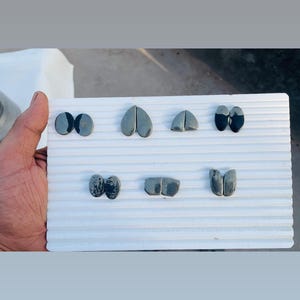 Puede incluir: Una colección de formas de piedra pulida de color gris oscuro, que incluye óvalos, corazones y otras formas abstractas, dispuestas sobre una superficie blanca y acanalada. Las piedras son sostenidas por una mano, mostrando su textura suave y diseños variados.
