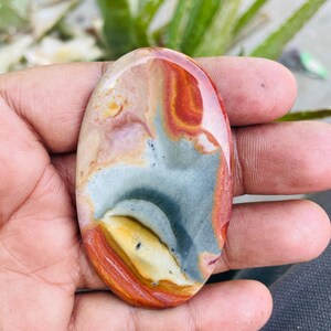 May include: An oval-shaped polished gemstone with a mix of colors. The stone features bands of red, orange, gray, and cream. The polished surface reflects light, highlighting the natural patterns and textures of the stone. A unique, natural stone.