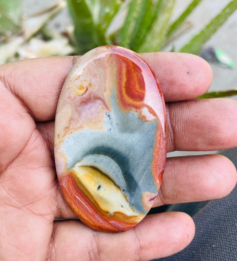 May include: An oval-shaped polished gemstone with a mix of colors. The stone features bands of red, orange, gray, and cream. The polished surface reflects light, highlighting the natural patterns and textures of the stone. A unique, natural stone.