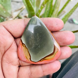 May include: A polished, teardrop-shaped gemstone with a mix of colors. The stone features a central area of muted green, bordered by bands of brown, orange, and a reddish-pink hue. The stone is held in a hand, with a blurred green plant in the background.