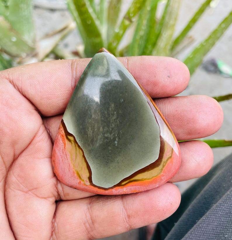 May include: A polished, teardrop-shaped gemstone with a mix of colors. The stone features a central area of muted green, bordered by bands of brown, orange, and a reddish-pink hue. The stone is held in a hand, with a blurred green plant in the background.