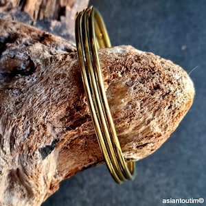 May include: A set of three gold-colored bangles, resting on a piece of weathered wood. The bangles are thin and round, reflecting light. The wood has a rough, textured surface with various shades of brown.