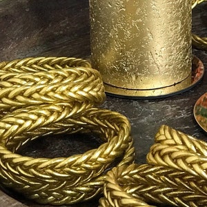 May include: A gold candle with a textured surface is lit and sitting on a wooden tray with gold rope trim.