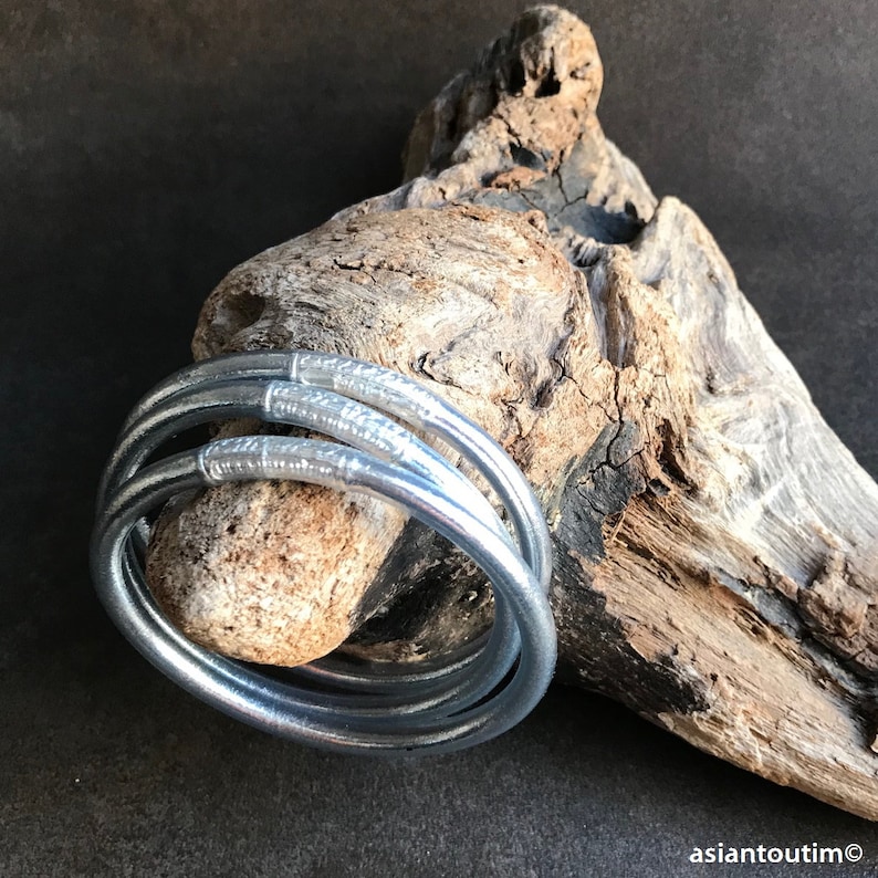 May include: Four silver bangles with a textured design. The bangles are stacked on top of each other and are resting on a piece of driftwood.