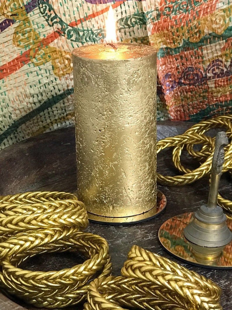 May include: A gold candle with a textured surface is lit and sitting on a wooden tray with gold rope trim.