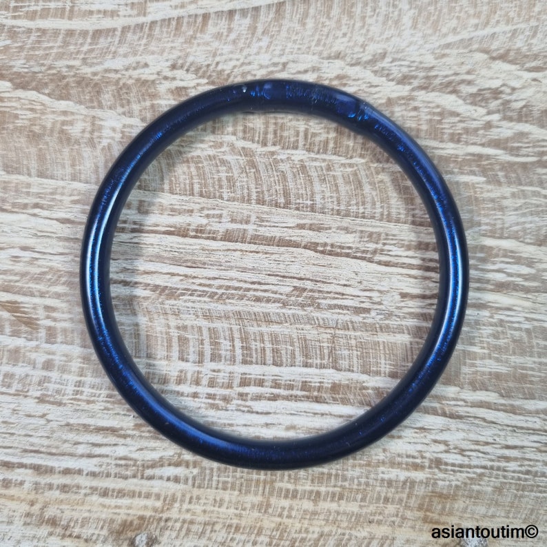 May include: A dark blue, circular bangle bracelet. The bracelet has a smooth, glossy finish and appears to be made of a solid material. The bangle is set against a light brown wooden surface.