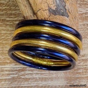May include: Stack of six bangles in alternating colors of gold and navy blue. The bangles are stacked on top of each other, with a wooden background. The bangles are smooth and shiny, reflecting light.