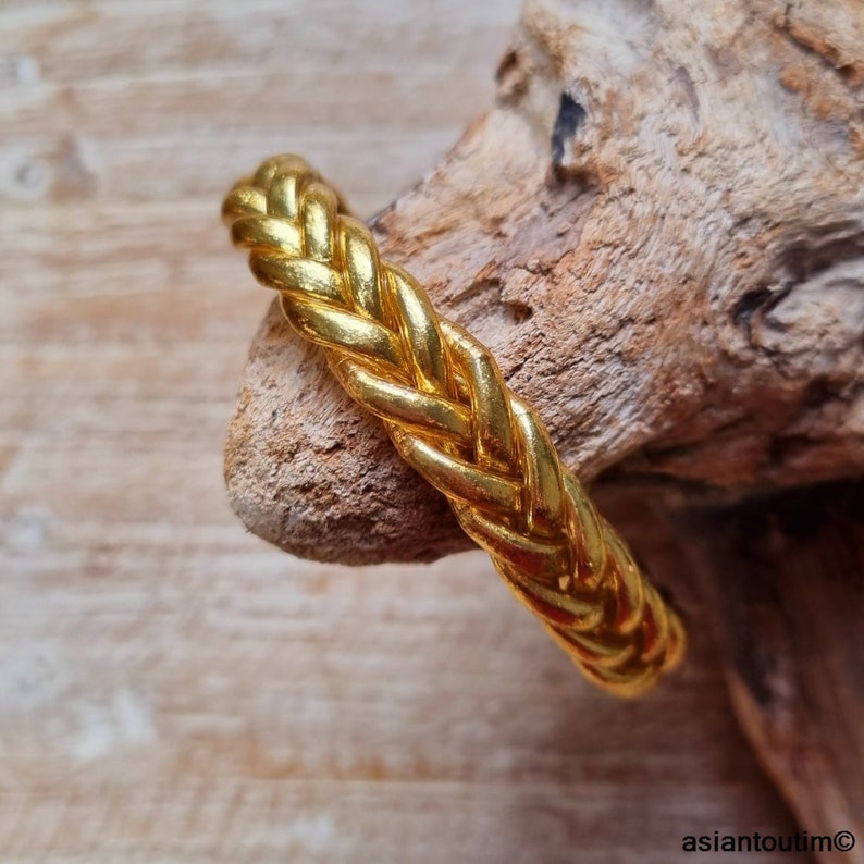 Puede incluir: Un brazalete trenzado de color dorado. El brazalete est&aacute; hecho de un metal grueso y tejido y tiene un acabado liso y pulido.