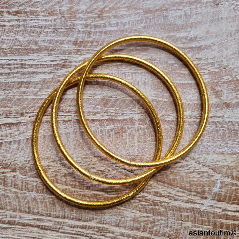 May include: Three gold-colored bangle bracelets stacked on top of each other. The bracelets are made of a woven material and have a shiny finish.