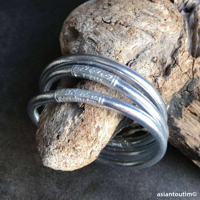 May include: Set of four silver bangles with decorative etched designs. The bangles are stacked on top of each other and are resting on a piece of driftwood.