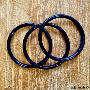 May include: Three dark blue bangles are intertwined on a light brown wooden surface. The bangles have a glossy finish and appear to be made of a solid material. The wooden surface has a natural grain pattern.