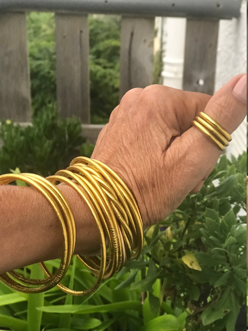 May include: A stack of gold-colored bangles on a person's wrist. The bangles are thin and have a slightly textured surface. There are also three gold-colored rings on the person's thumb.