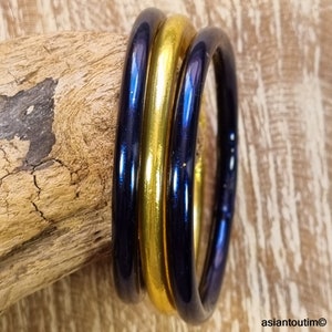 May include: Three glass bangles: two dark blue and one gold. The bangles are stacked together, reflecting light. The background is a piece of wood with a rough texture. The bangles are a simple, elegant accessory.