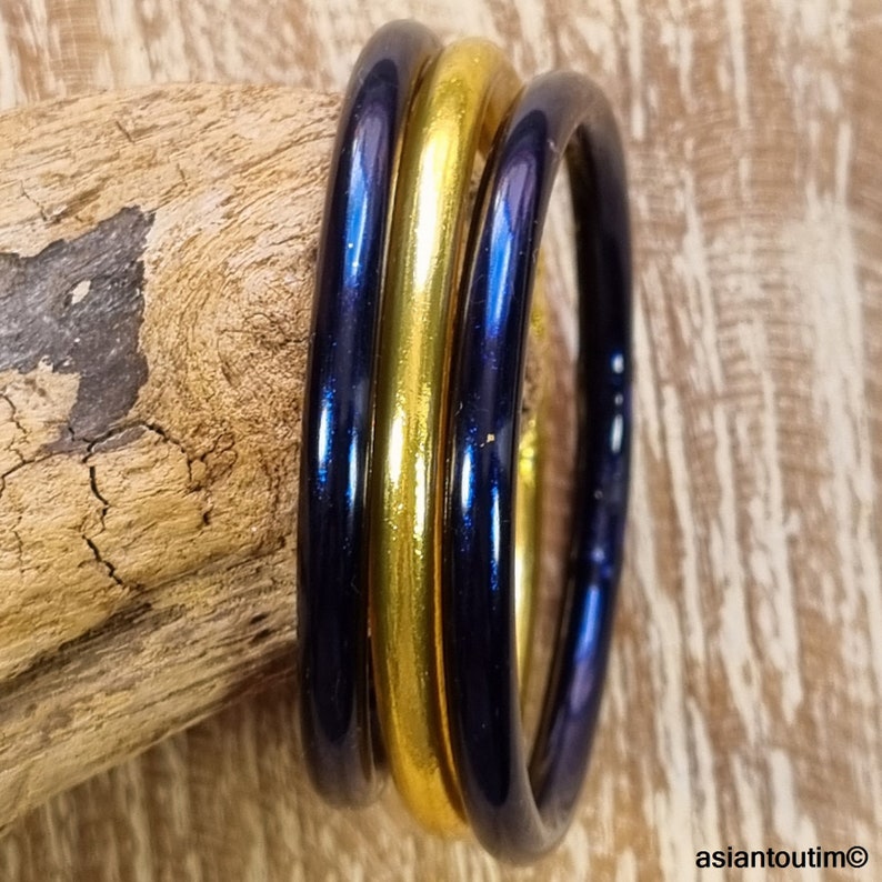 May include: Three glass bangles: two dark blue and one gold. The bangles are stacked together, reflecting light. The background is a piece of wood with a rough texture. The bangles are a simple, elegant accessory.