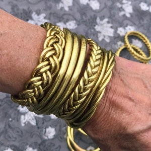 Puede incluir: Un conjunto de pulseras de metal dorado, algunas con un dise&ntilde;o trenzado, apiladas en la mu&ntilde;eca de una persona.