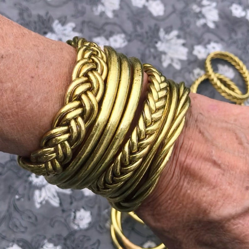Puede incluir: Un conjunto de pulseras de metal dorado, algunas con un dise&ntilde;o trenzado, apiladas en la mu&ntilde;eca de una persona.