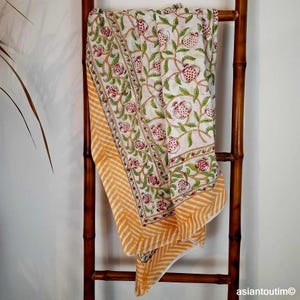Peut inclure: Textile blanc et crème avec un motif de grenade et de feuilles, avec des accents rouges et verts. La bordure présente un motif en chevrons orange et blanc. Le textile est drapé sur une échelle en bambou.