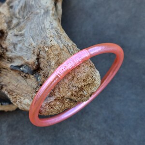 K&ouml;nnte beinhalten: Ein durchscheinendes, korallenfarbenes Armreif-Armband mit dezentem Schimmer. Das Armband liegt auf einem St&uuml;ck verwittertem Holz und zeigt seine glatte, abgerundete Form. Das Armband hat einen dekorativen Abschnitt mit ge&auml;tzten Details.
