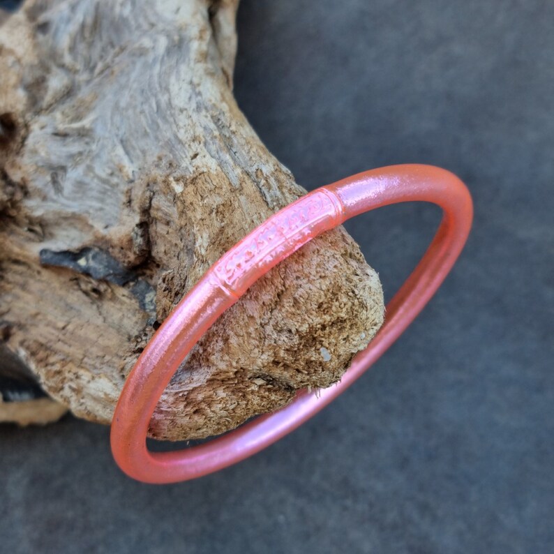 K&ouml;nnte beinhalten: Ein durchscheinendes, korallenfarbenes Armreif-Armband mit dezentem Schimmer. Das Armband liegt auf einem St&uuml;ck verwittertem Holz und zeigt seine glatte, abgerundete Form. Das Armband hat einen dekorativen Abschnitt mit ge&auml;tzten Details.