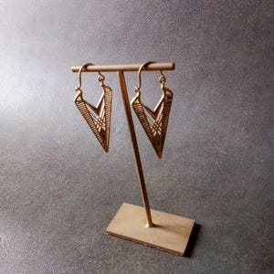 May include: A pair of gold-colored earrings with a geometric arrow-like design. The earrings are displayed on a gold-colored stand against a gray background. The earrings have a triangular shape with intricate line details.