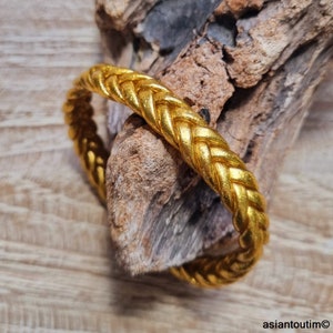 May include: A gold-colored braided bangle bracelet. The bracelet is made of a thick, woven metal and has a smooth, polished finish.