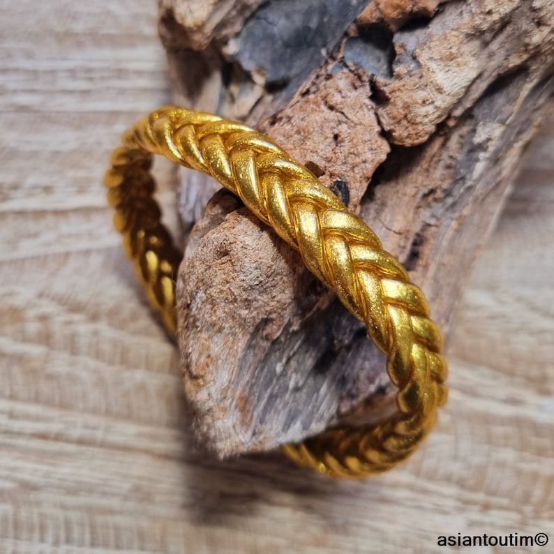 May include: A gold-colored braided bangle bracelet. The bracelet is made of a thick, woven metal and has a smooth, polished finish.