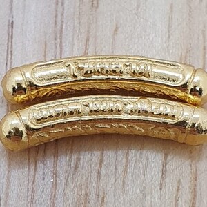 May include: Two gold-colored metal charms with intricate designs and Thai script. The charms are curved and have rounded ends.