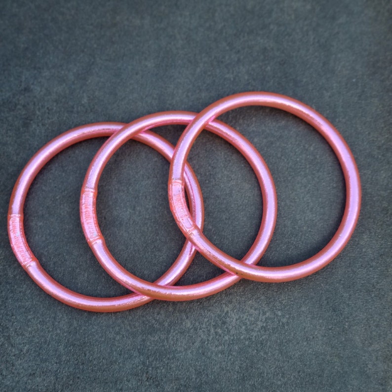 K&ouml;nnte beinhalten: Drei rosa, kreisf&ouml;rmige Armreifen mit dezentem Schimmer liegen auf einer dunkelgrauen Oberfl&auml;che. Die Armreifen haben ein glattes, abgerundetes Design mit einem leicht strukturierten Abschnitt. Die Gesamt&auml;sthetik ist schlicht und elegant.