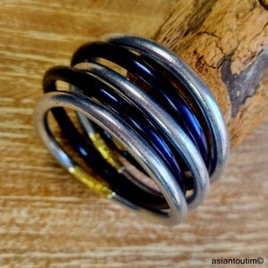 May include: A stack of five rings in silver, black, and blue. The rings are smooth and metallic, with a slight sheen. The background is a light brown wooden surface.