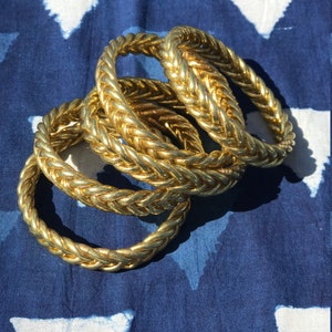 Puede incluir: Cuatro pulseras doradas trenzadas apiladas unas sobre otras sobre una tela de patr&oacute;n azul y blanco.