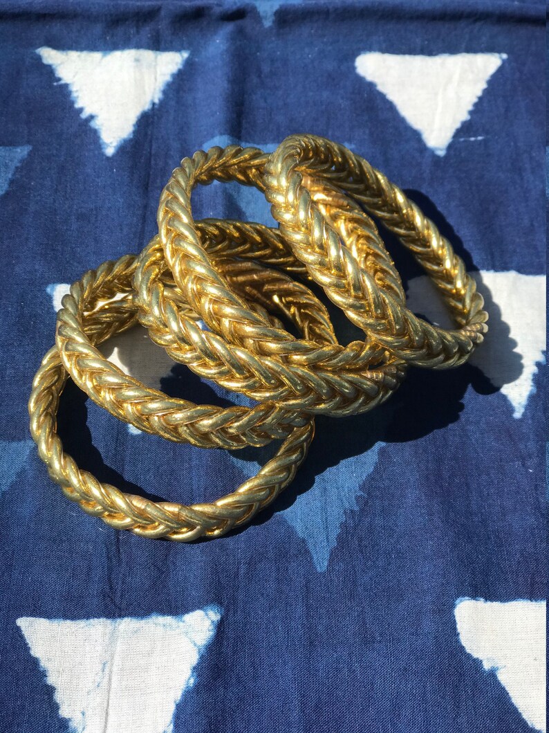 Puede incluir: Cuatro pulseras doradas trenzadas apiladas unas sobre otras sobre una tela de patr&oacute;n azul y blanco.