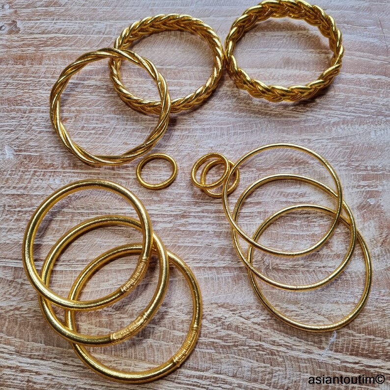 Puede incluir: Un conjunto de diez brazaletes de color dorado, algunos con un dise&ntilde;o trenzado, dispuestos sobre una superficie de madera clara.